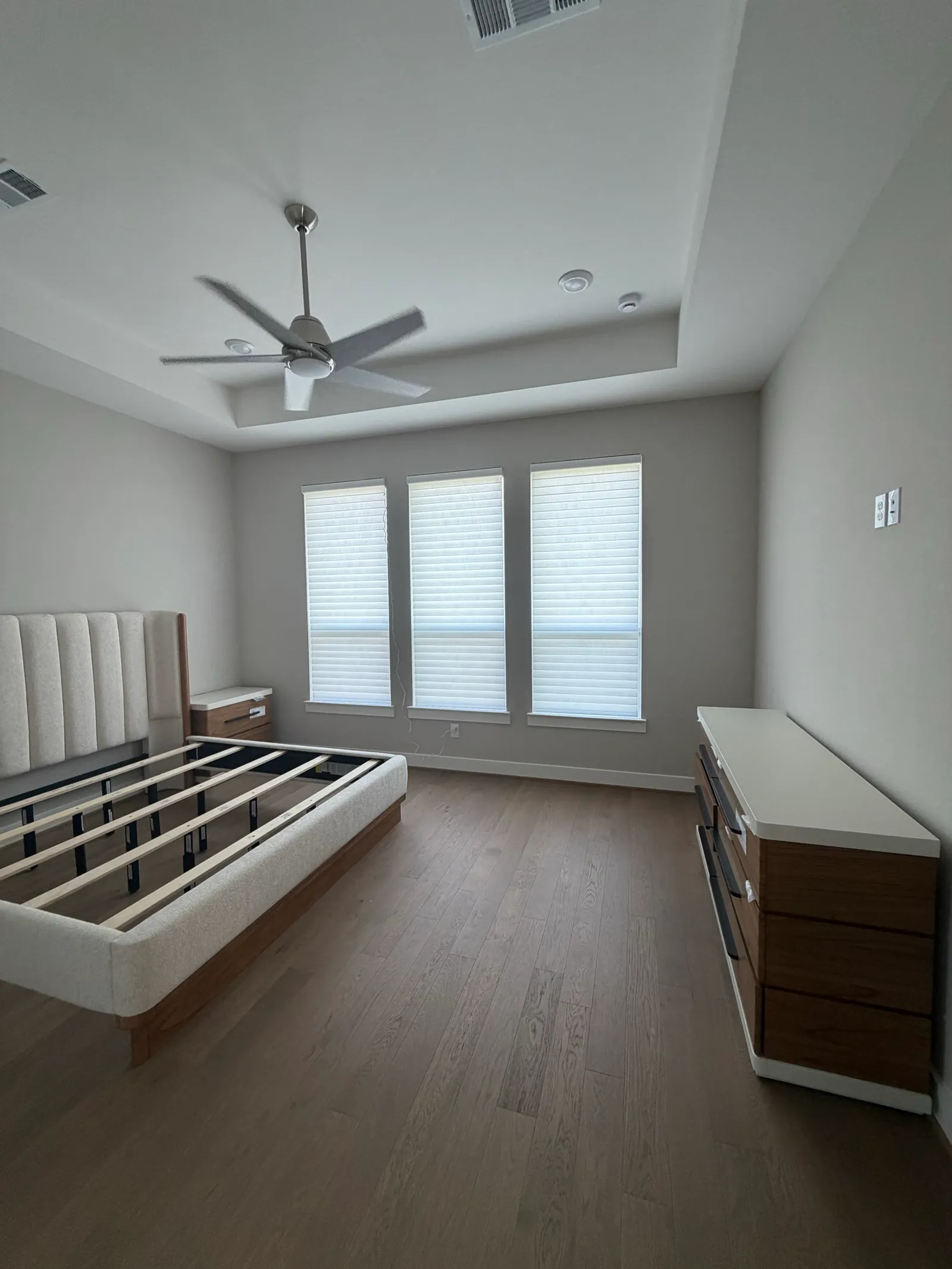 Wide bedroom view with three white sheer shades, a wood bed frame and ceiling fan.