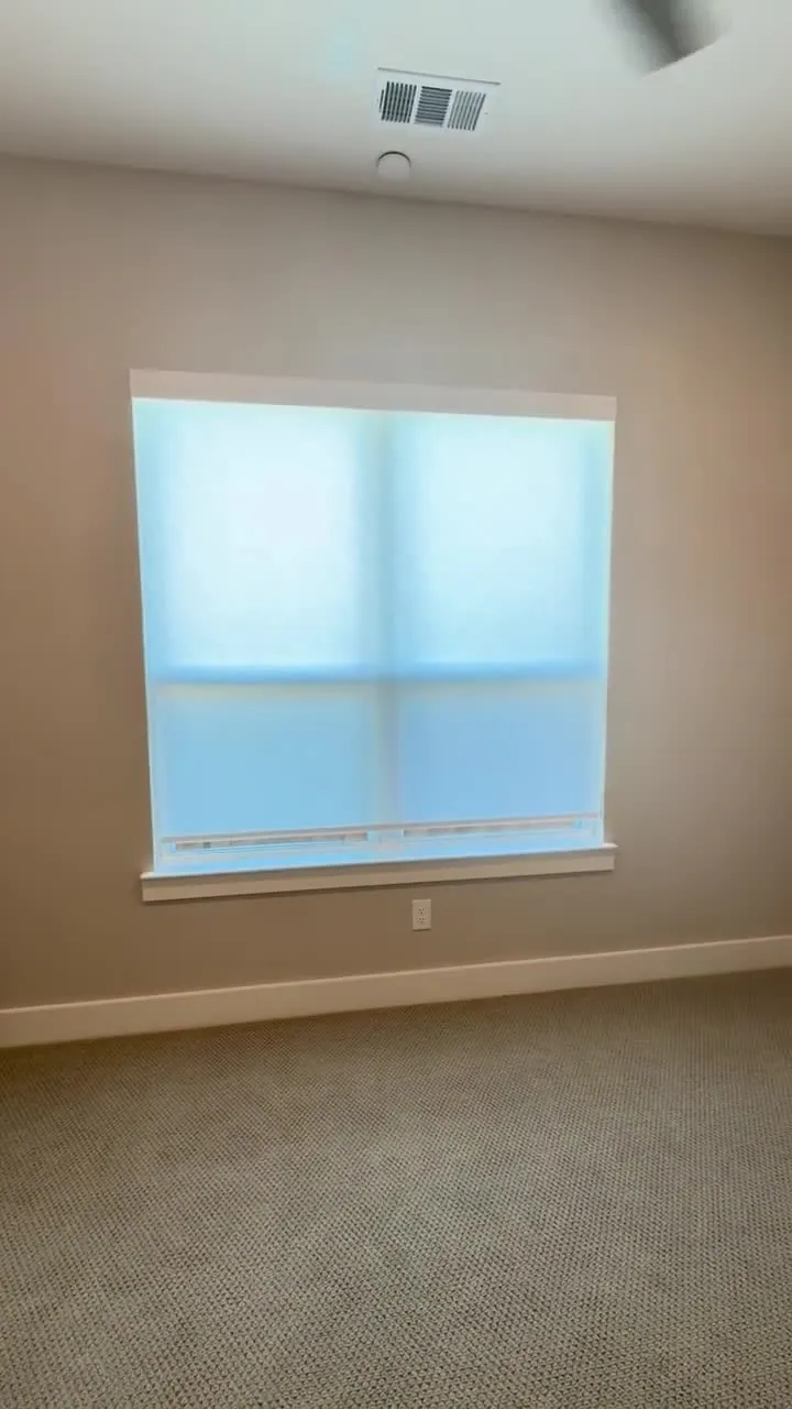 White roller shade lowering over a window in a carpeted room.